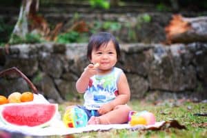 Baby girl sitting on blanket feeding herself picnic foods